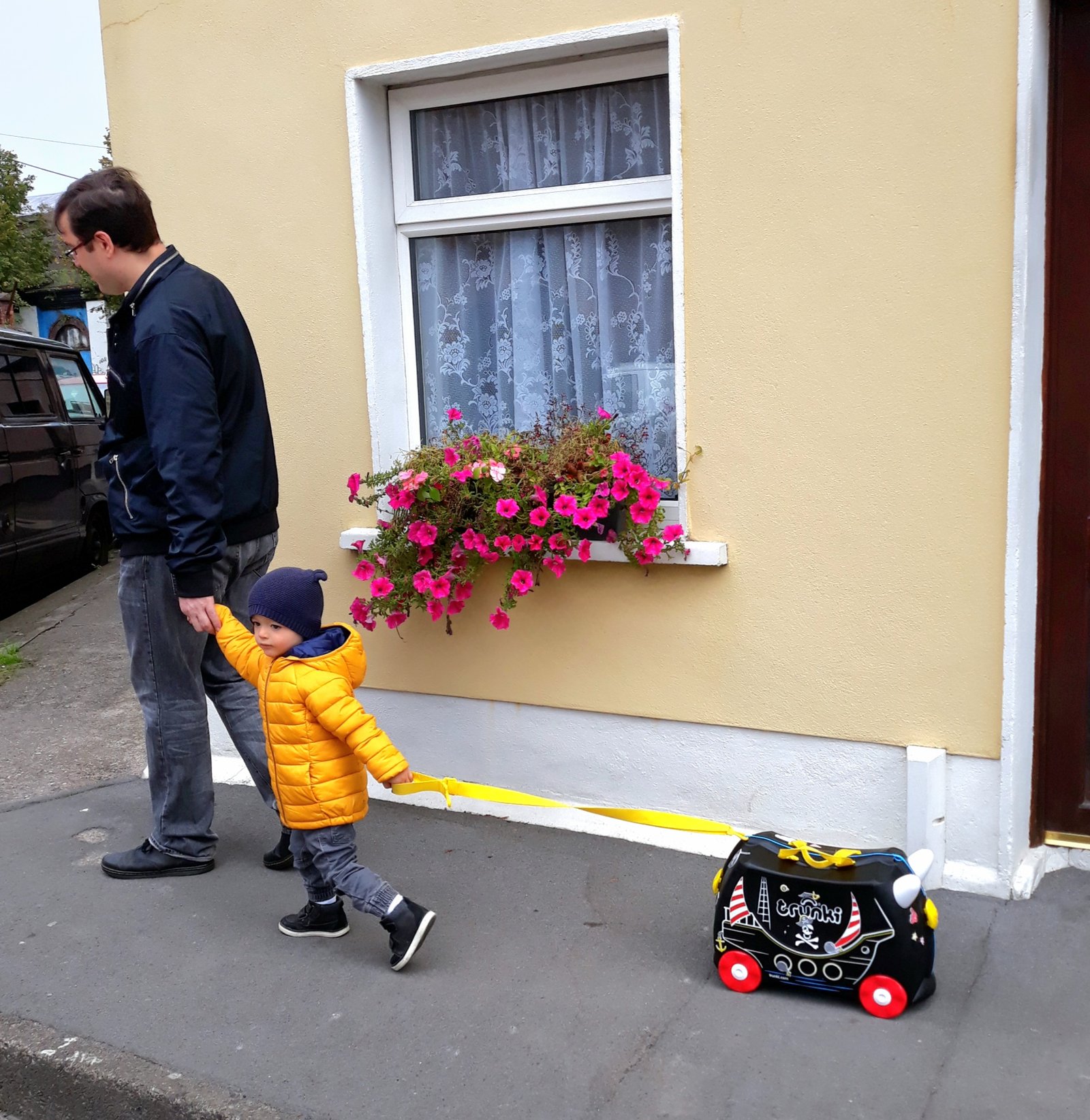 Our young son pulling his pirate-ship Trunki ride-on suitcase along a street past a flower-filled window box, walking hand in hand with his dad