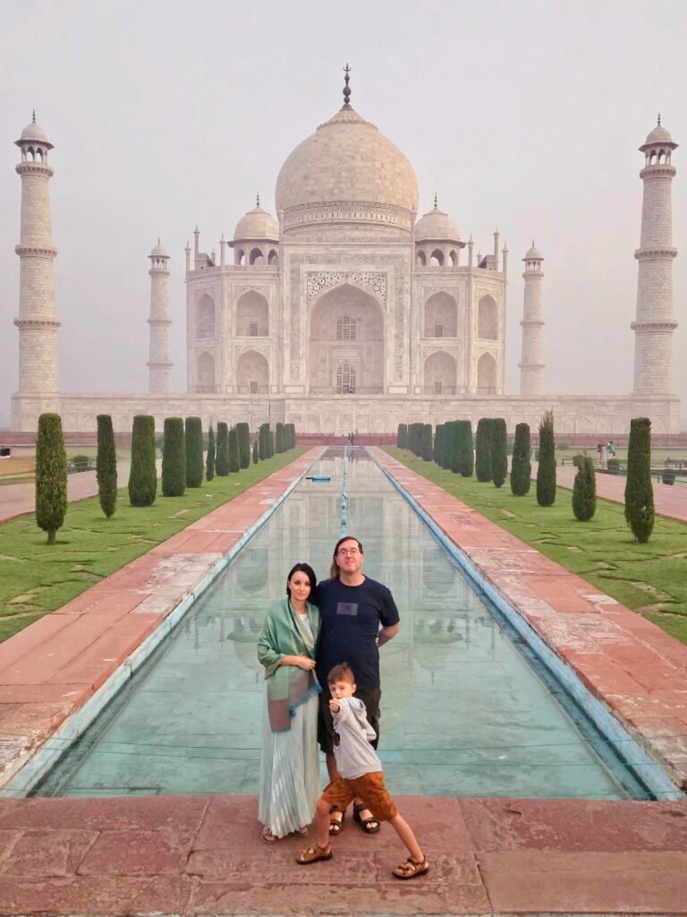 Our worldschooling family of three standing in front of the Taj Mahal in Agra, India, beside the reflecting pool on a misty morning