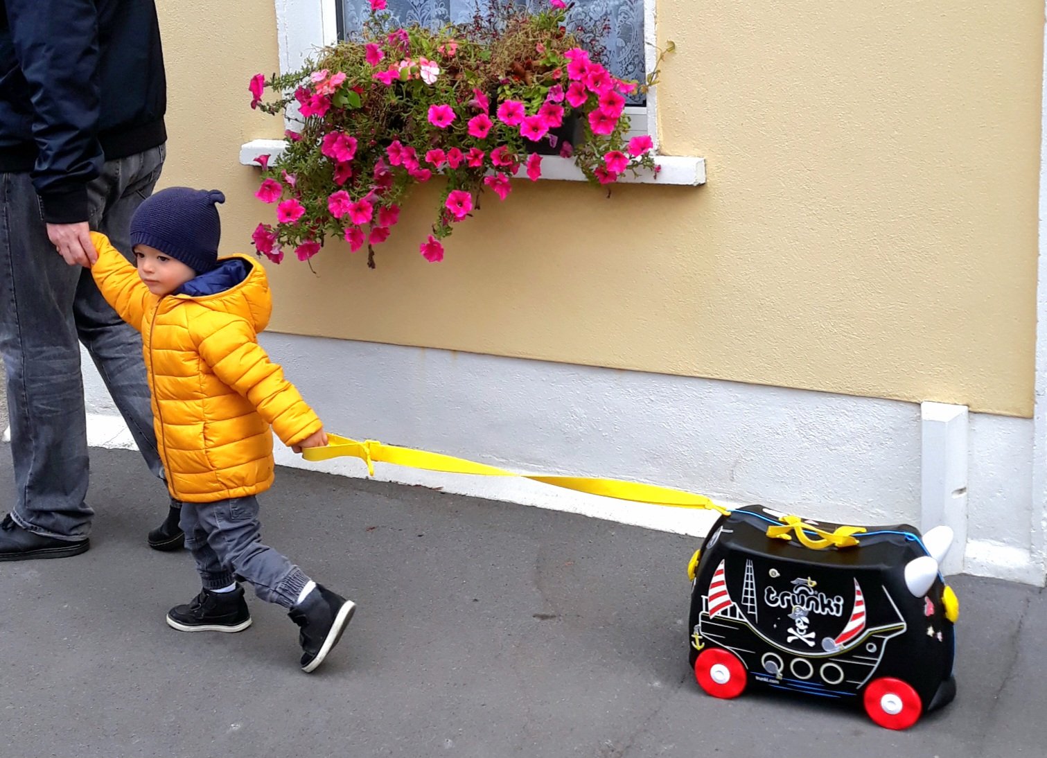 Our young son pulling his pirate-ship Trunki ride-on suitcase along a street past a flower-filled window box, walking hand in hand with his dad