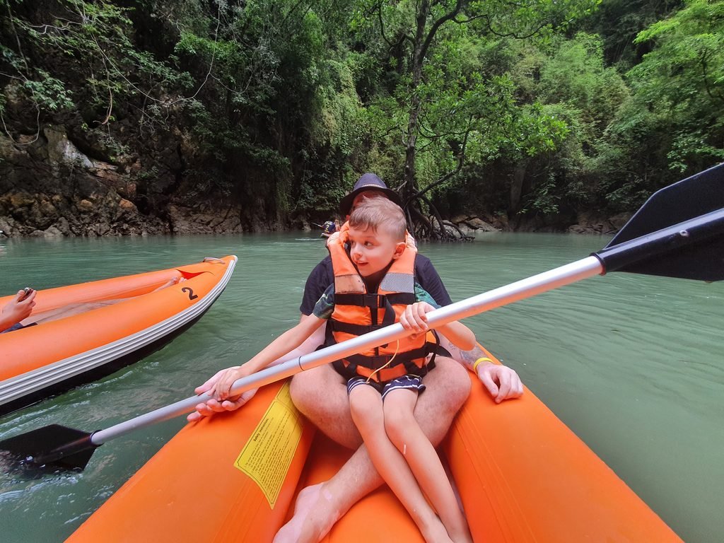 Boy in a life jacket sharing an inflatable kayak with a parent on calm green water in Thailand